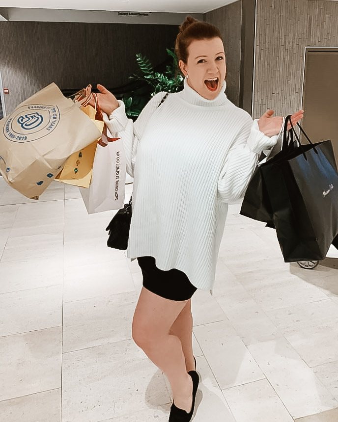 A woman in white jumper holding up shopping bags smiling